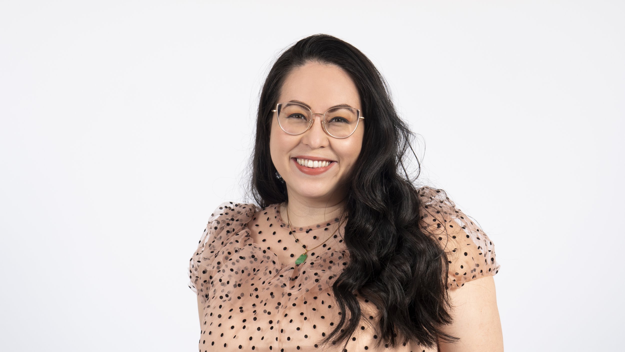 A woman with long dark wavy hair, wearing glasses, a pink polka dot top, and a green pendant necklace, smiling at the camera against a plain white background.