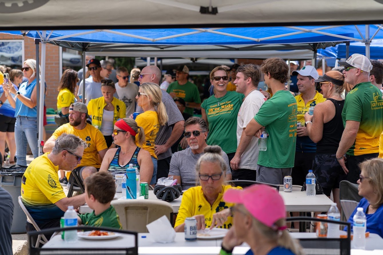 Group of people at an outdoor event, some wearing yellow and green shirts, under tents. Participants are socializing and eating at tables with drinks and snacks visible.