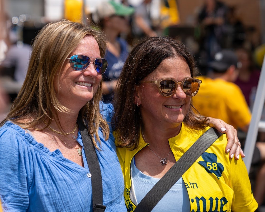 Two women smiling and posing outdoors, wearing sunglasses. One is in a blue shirt, the other in a yellow jacket with a number and flower emblem. The background shows a busy, sunlit environment with people.
