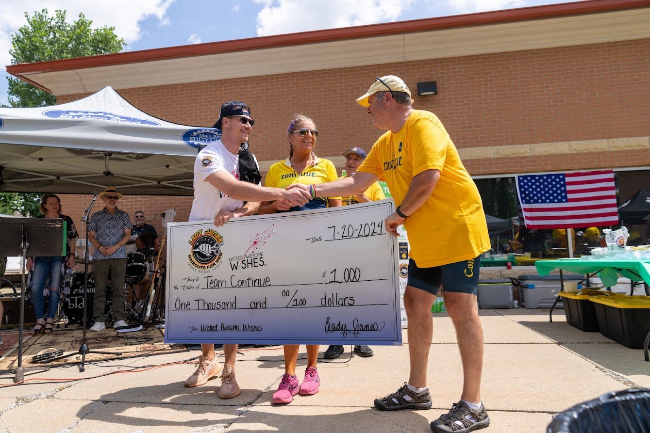 People at an outdoor event holding a large ceremonial check for $1,000 made out to "Team Continue" dated July 20, 2024, from "Wicked Awesome Wishes." The background shows a band and an American flag.