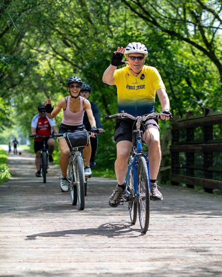 Group of cyclists riding on a sunny wooded path, smiling and waving.