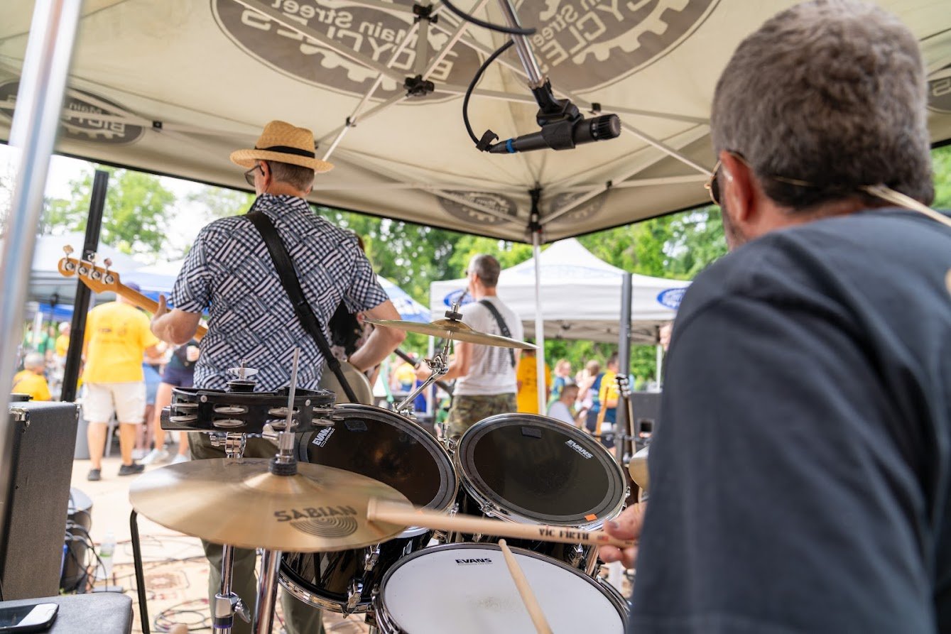 View from behind an outdoor band setup with a drummer and guitarist playing under a canopy, audience in the background.