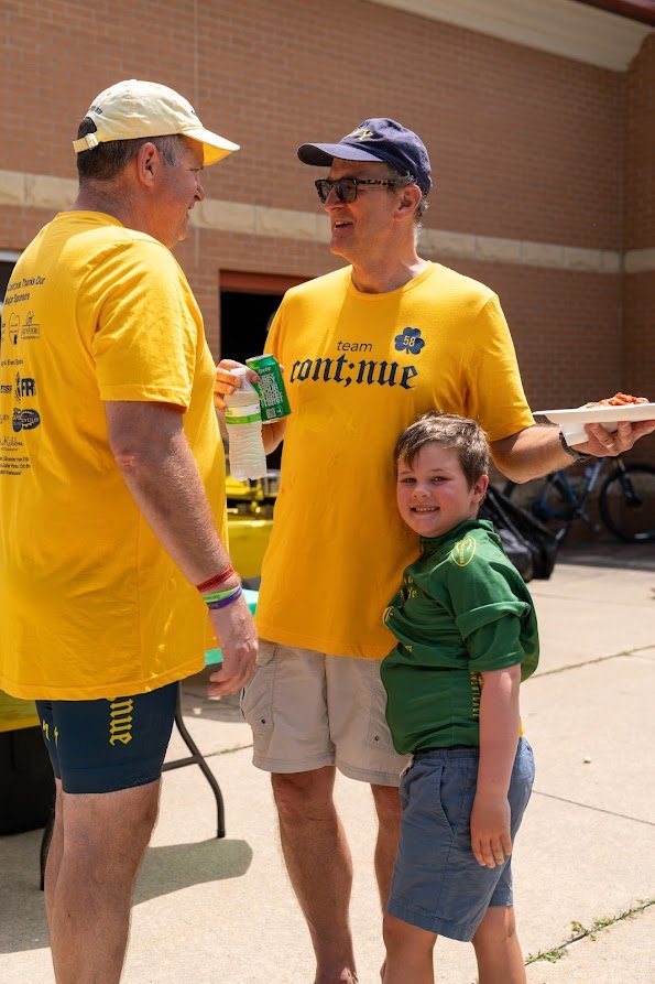 Two men wearing yellow "team continue" shirts and a child standing outdoors, with one man holding a plate of food and a can, smiling and talking.
