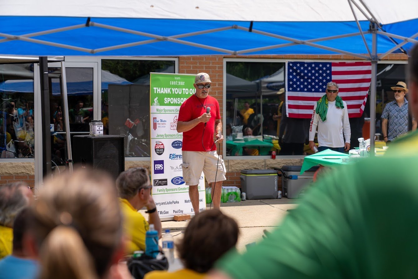 Outdoor event with a man in a red shirt speaking under a canopy, surrounded by people, a sponsor board, and an American flag in the background.