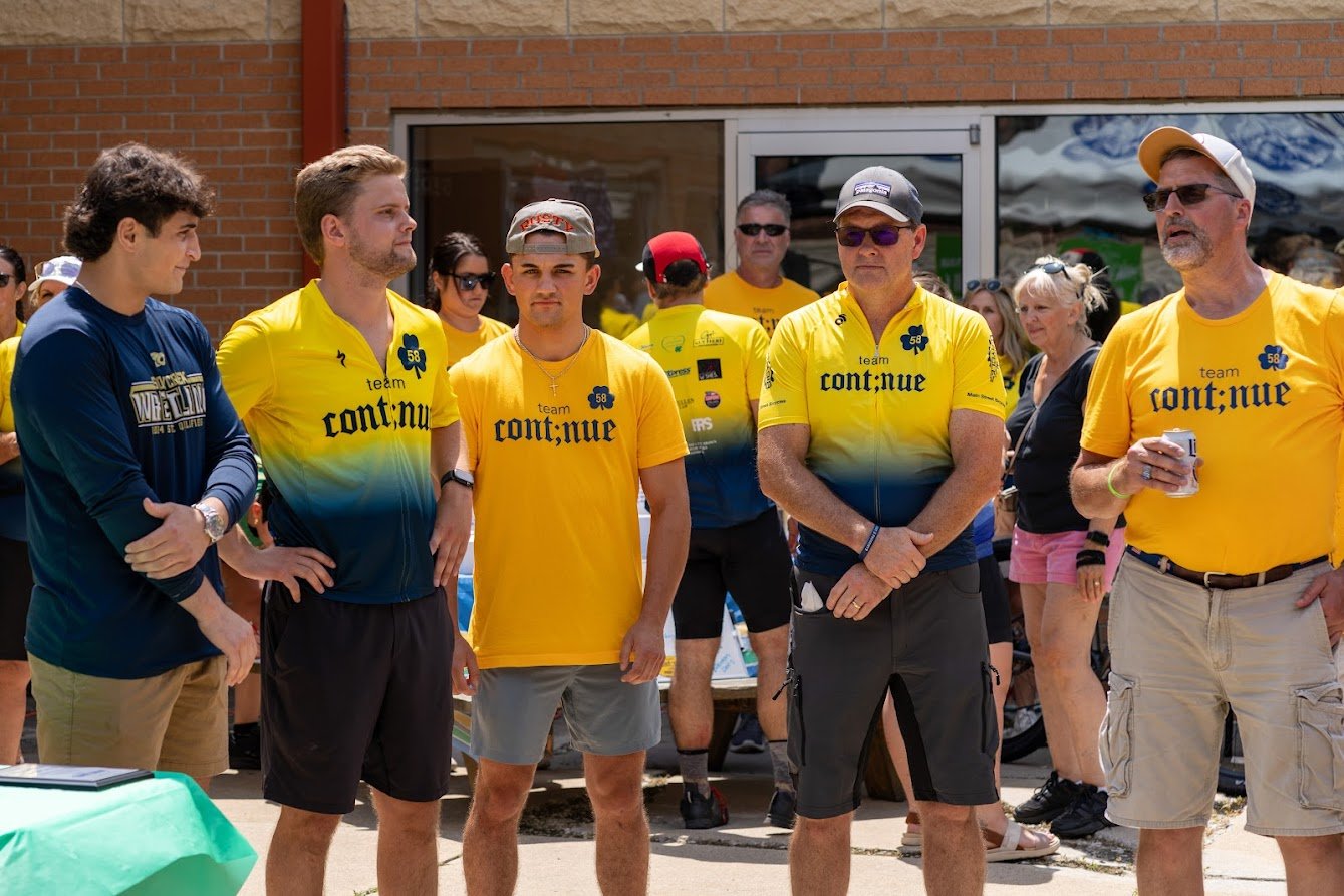 Group of men wearing yellow and blue team shirts standing outdoors in front of a crowd.
