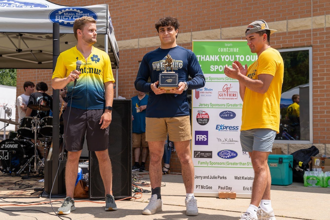 Three young men stand outdoors at an event. The first man holds a microphone, the second man holds a trophy, and the third is clapping. They are beside a banner displaying sponsor logos. In the background, there is a tent and a musician near a drum s