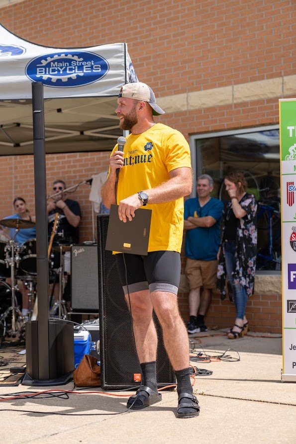 Man in yellow shirt holding a microphone speaks at an outdoor event with a band and people in the background.