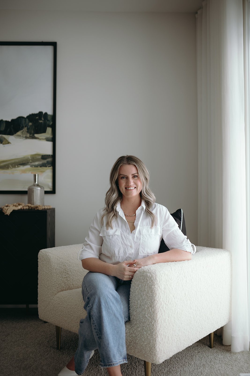 A woman with blonde hair, wearing a white button-up shirt and jeans, sitting on a cream-colored chair, smiling. There is a black sideboard with a decorative bottle and a painting on the wall behind her, with a window with white curtains to her right.
