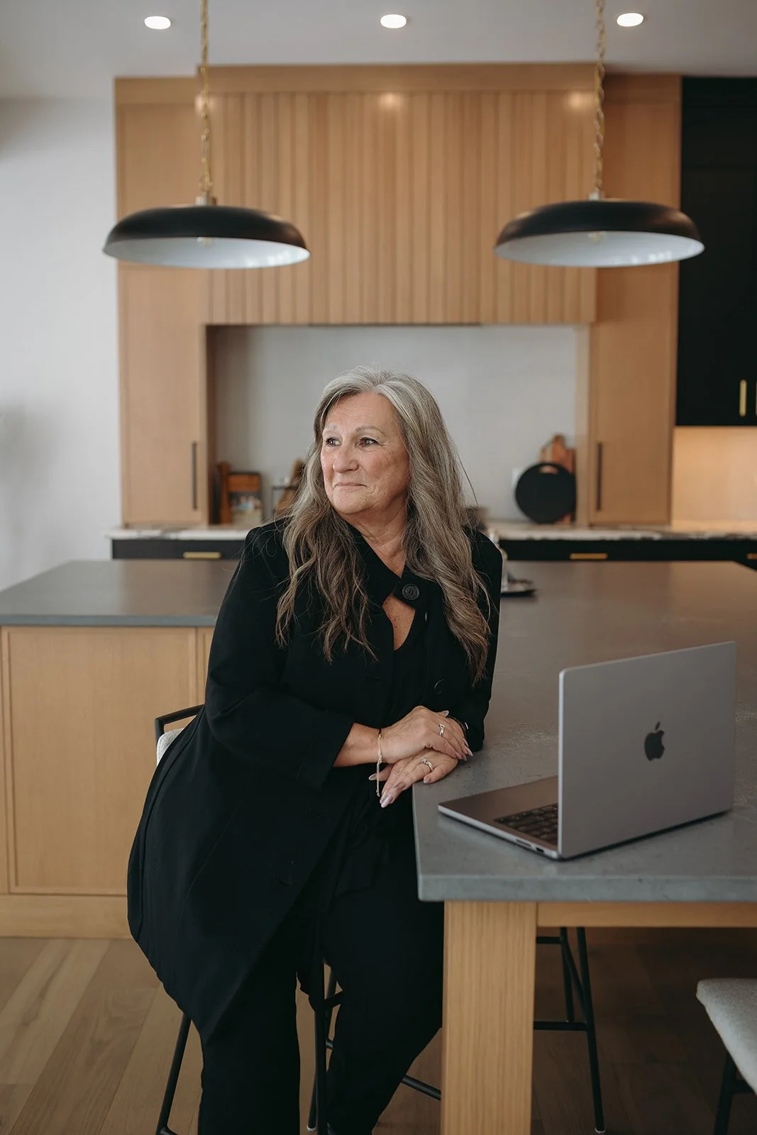 An older woman with gray hair sitting at a kitchen island with a laptop in front of her, wearing a black coat.