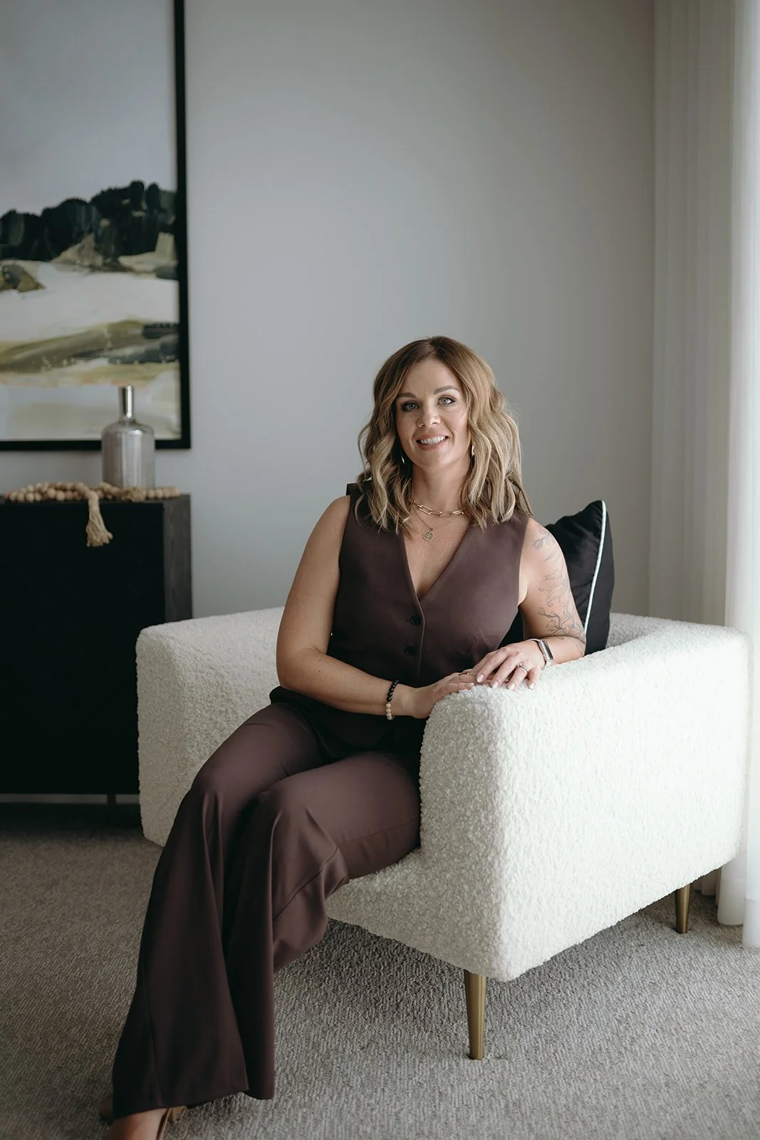 A woman sitting on a white textured armchair with a black pillow, wearing a brown sleeveless top and matching pants, smiling, with artwork and decor on the wall behind her.