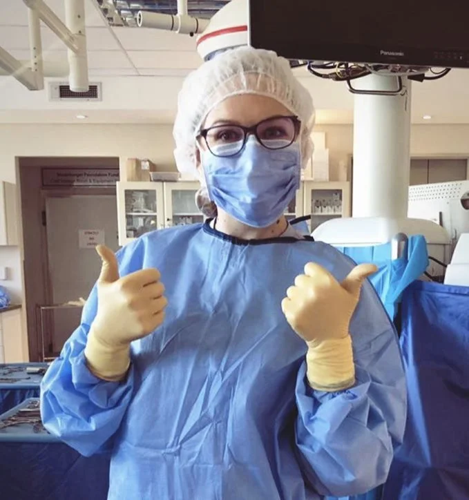 Healthcare professional in scrubs, mask, and gloves giving a thumbs-up gesture.