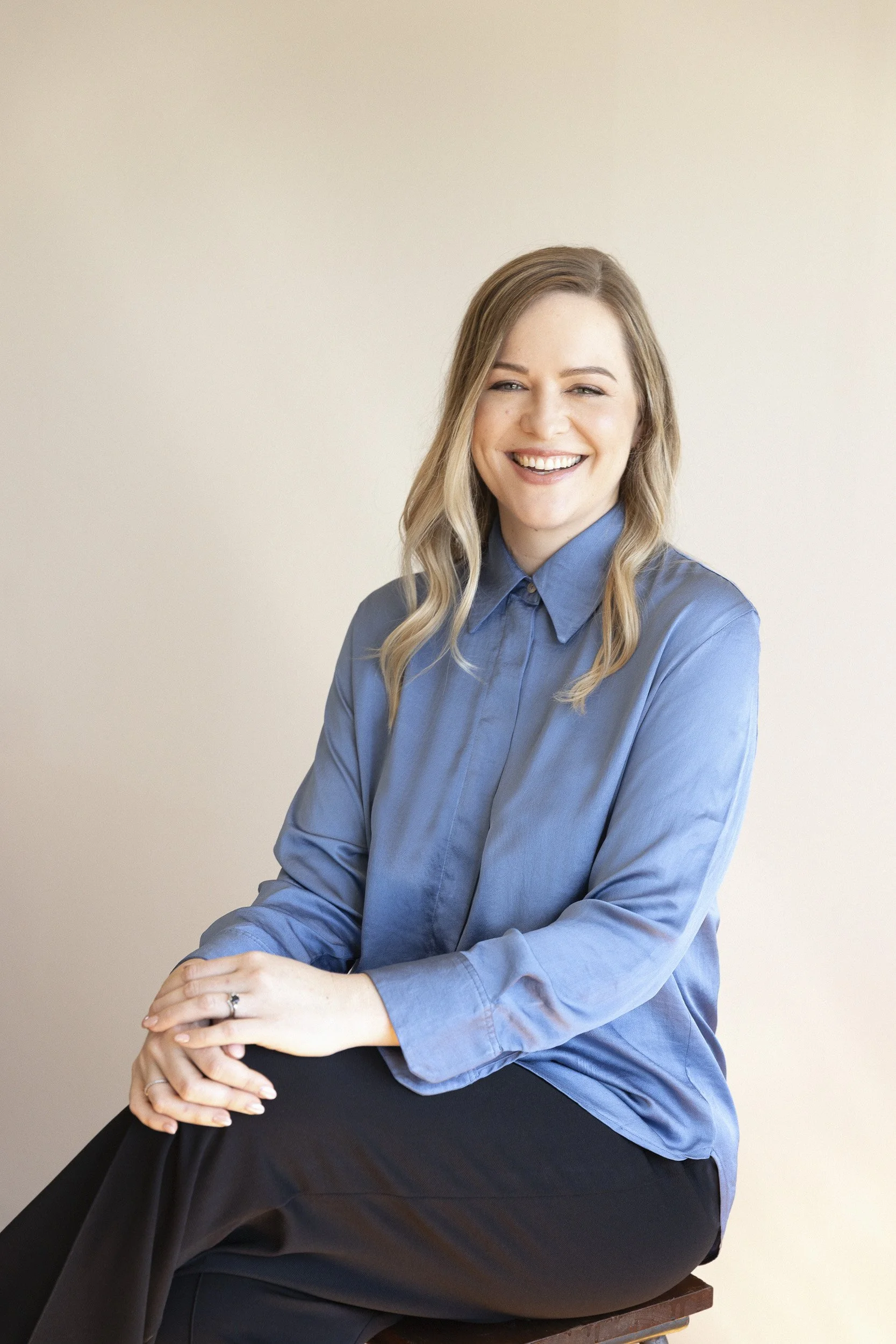 Portrait of a smiling woman with blonde wavy hair, wearing a blue button-up shirt and black pants, sitting on a stool against a plain light-colored wall.