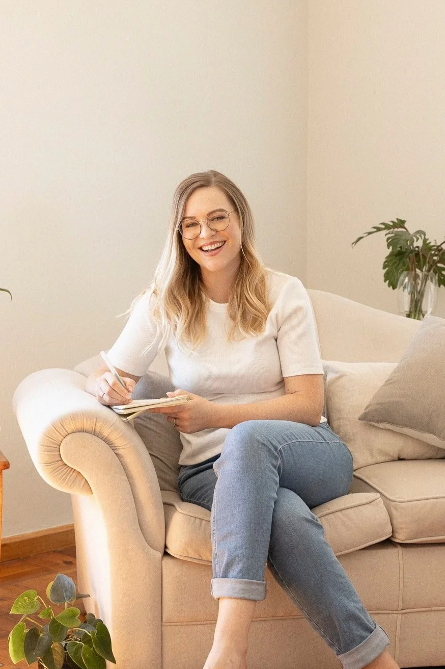 A woman with blonde hair and glasses sitting on a beige sofa, smiling and writing in a notebook. She is wearing a white t-shirt and rolled-up jeans.