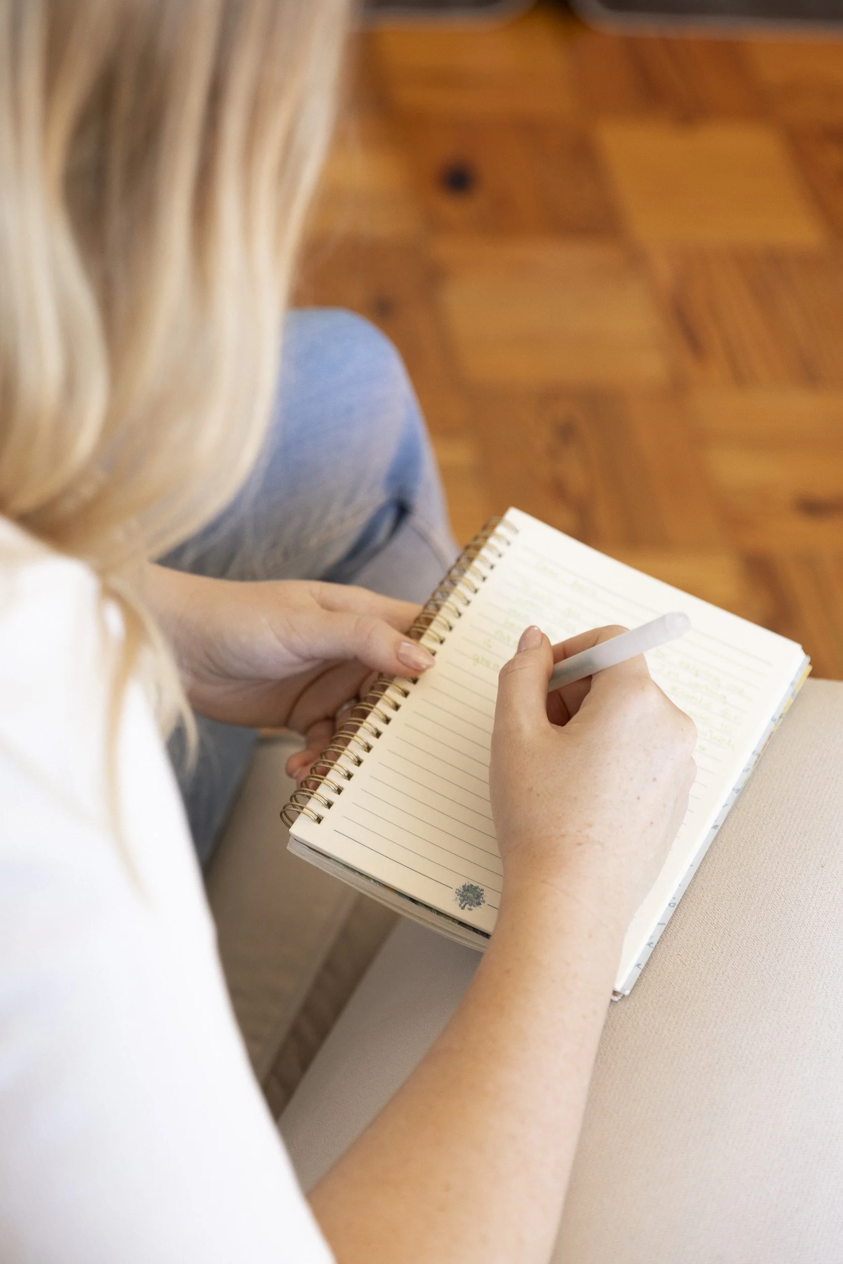 A woman with blonde hair taking notes in a lined notebook with a white pen, sitting on a beige chair near a hardwood floor.