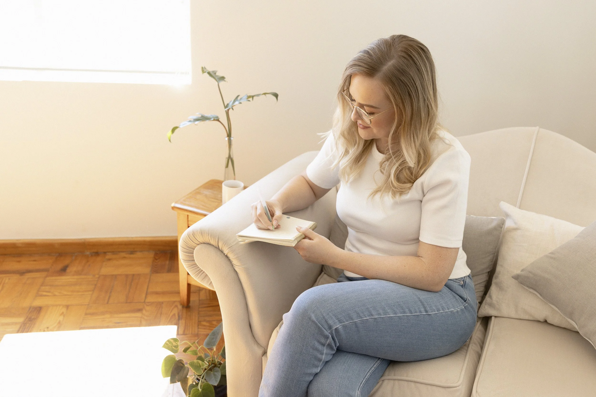 A woman with blonde hair and glasses sitting on a beige couch, writing in a notebook with a pen in a softly lit living room.