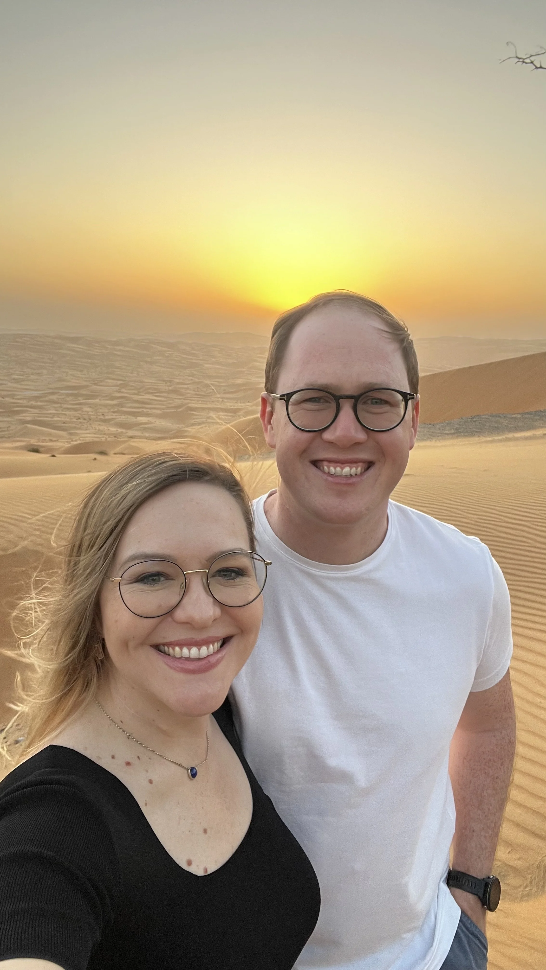 A smiling couple taking a selfie in a desert at sunset, with dunes in the background.