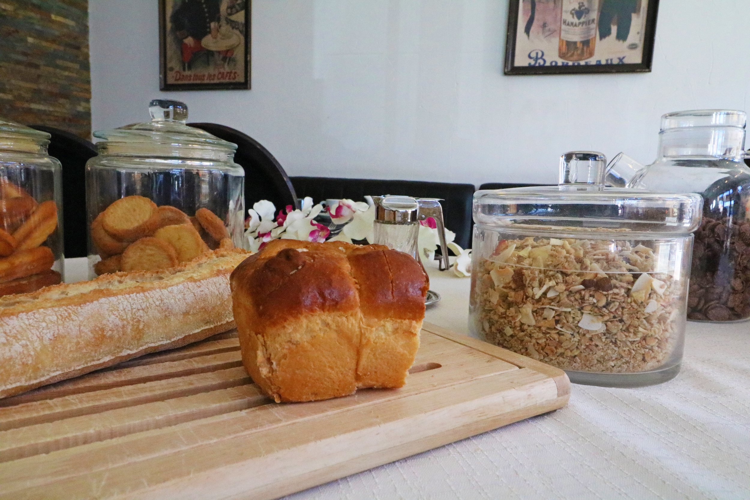 Petit-déjeuner avec pain, brioche, biscuits, et granola dans des bocaux en verre sur une table en bois.