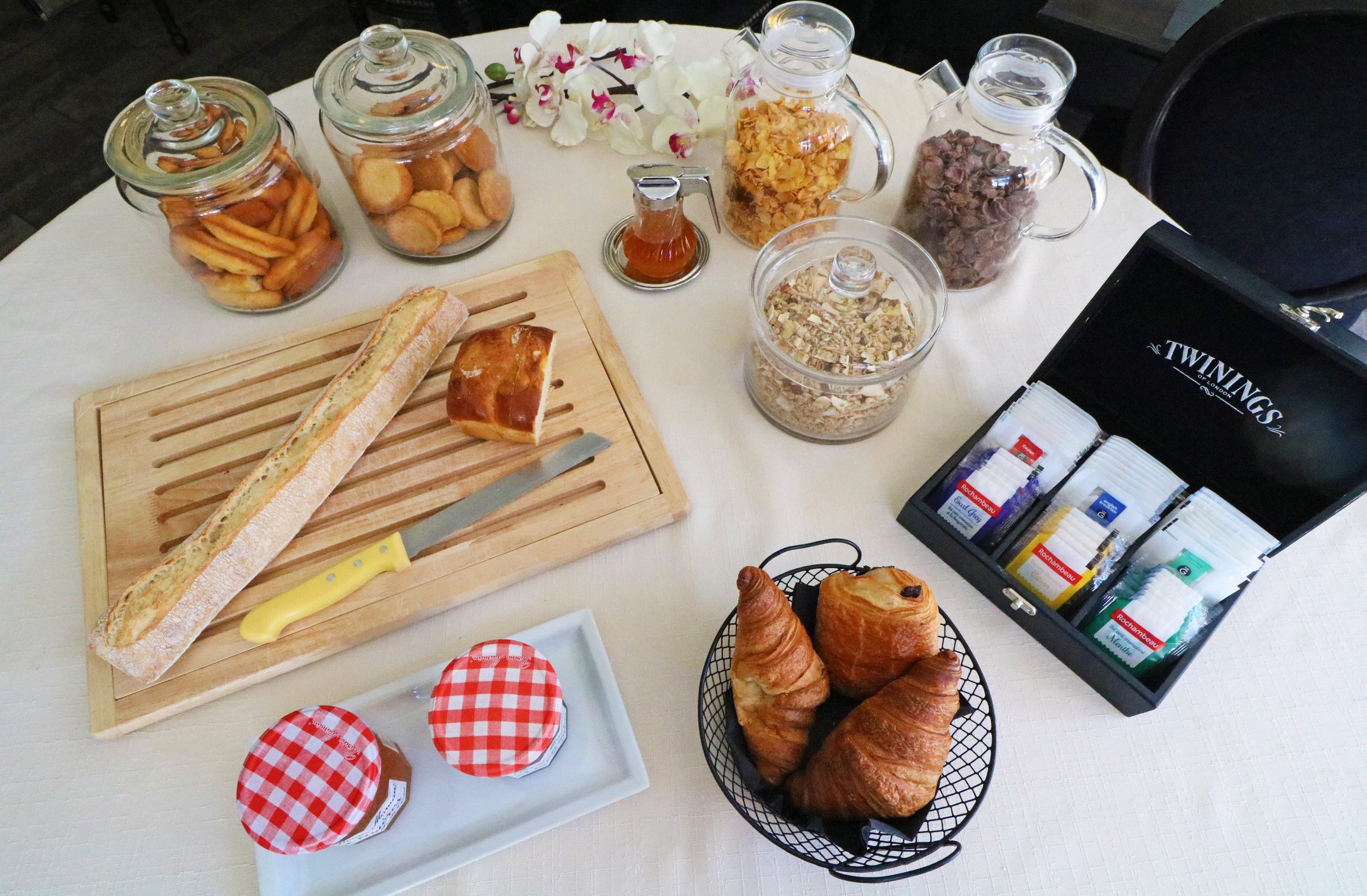 Petit-déjeuner avec baguette, couteau, confiture, croissants, viennoiseries, pots de céréales et biscuits, boîte de sachets de thé, et fleur d'orchidée sur une table blanche.