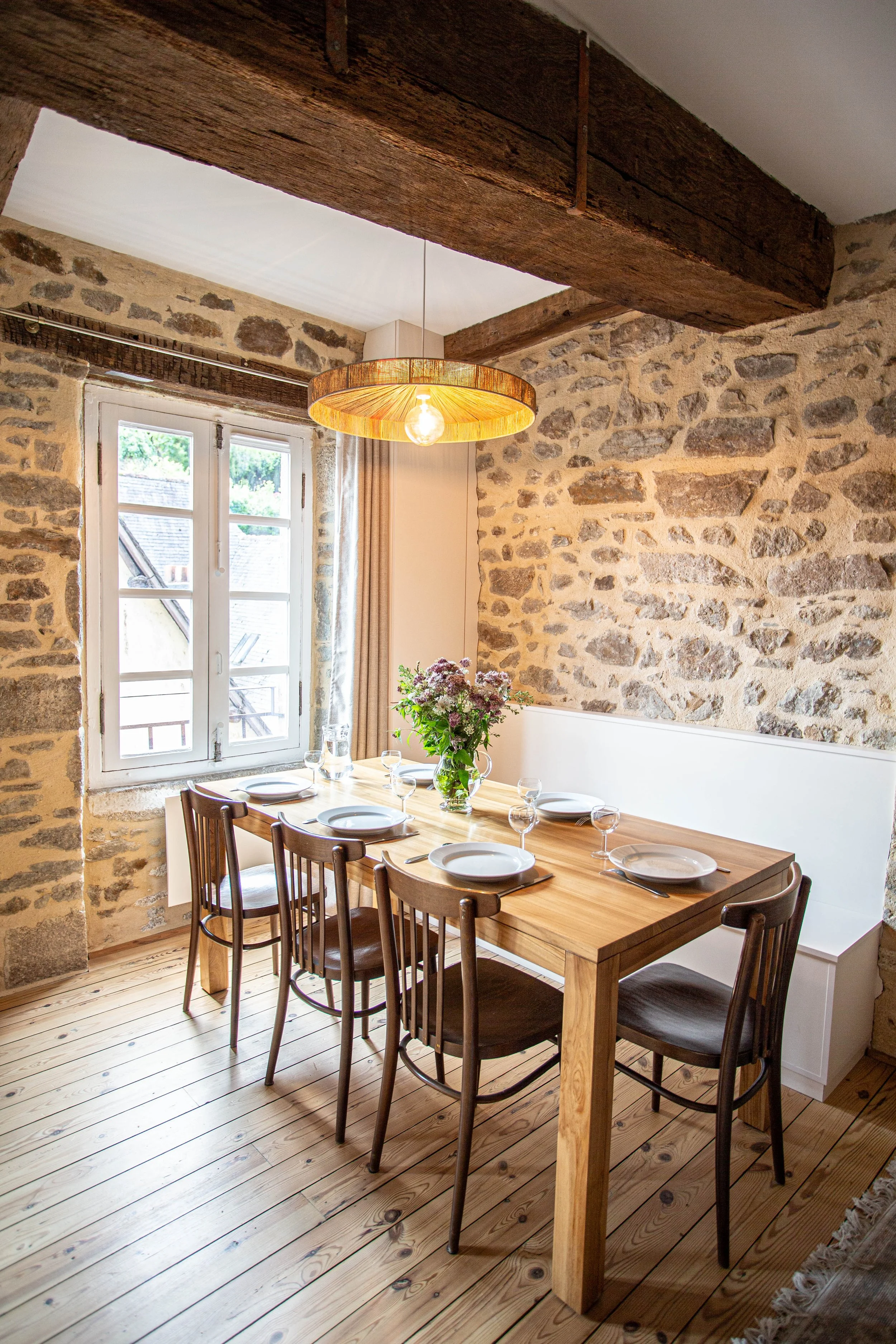 Salle à manger avec murs en pierres, fenêtre, table en bois avec six chaises, vaisselle et fleurs, plafond avec poutres en bois et luminaire