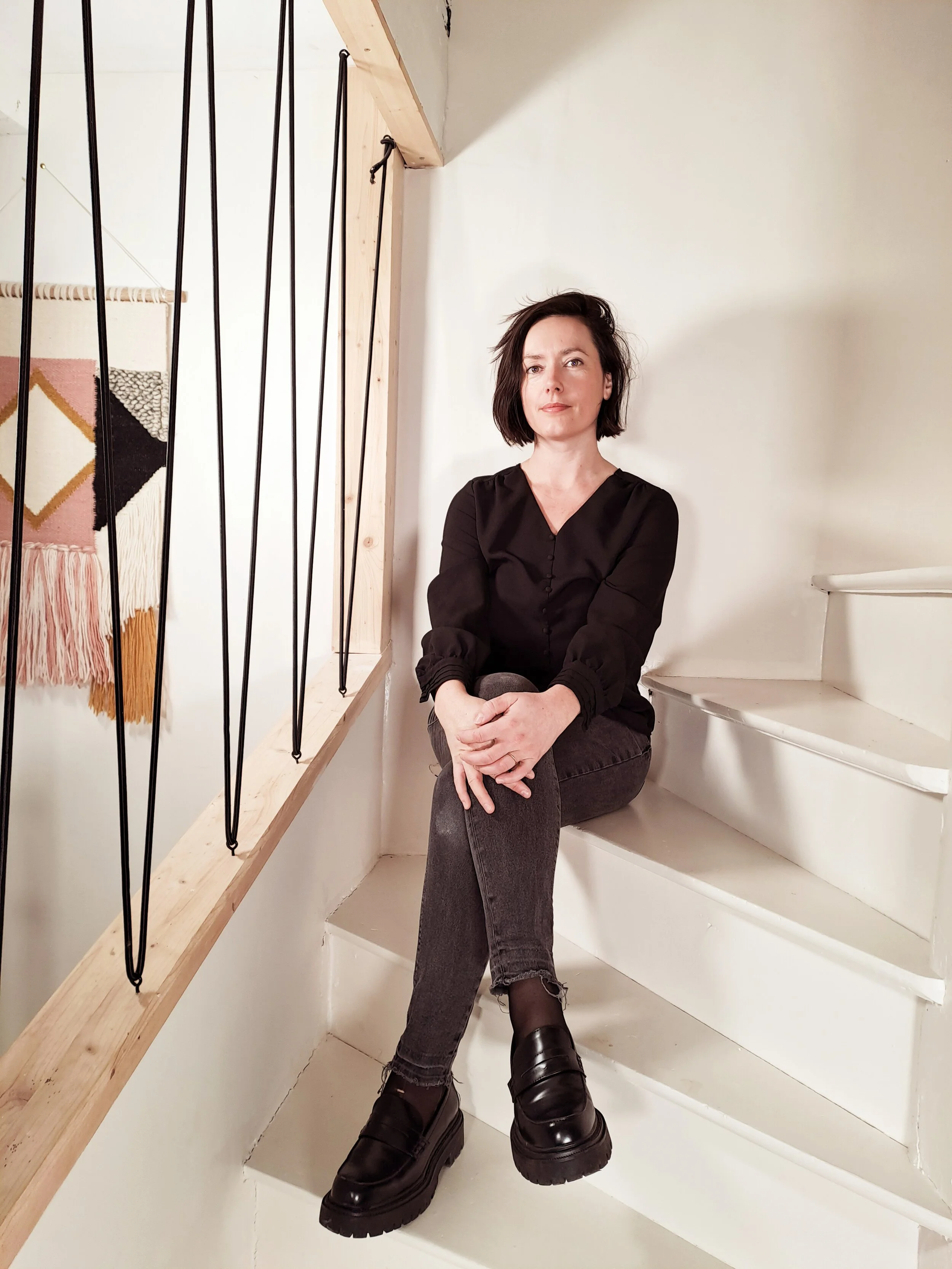 Une femme assise sur un escalier blanc en intérieur, portant un haut noir, un jean gris déchiré et des chaussures noires robustes.