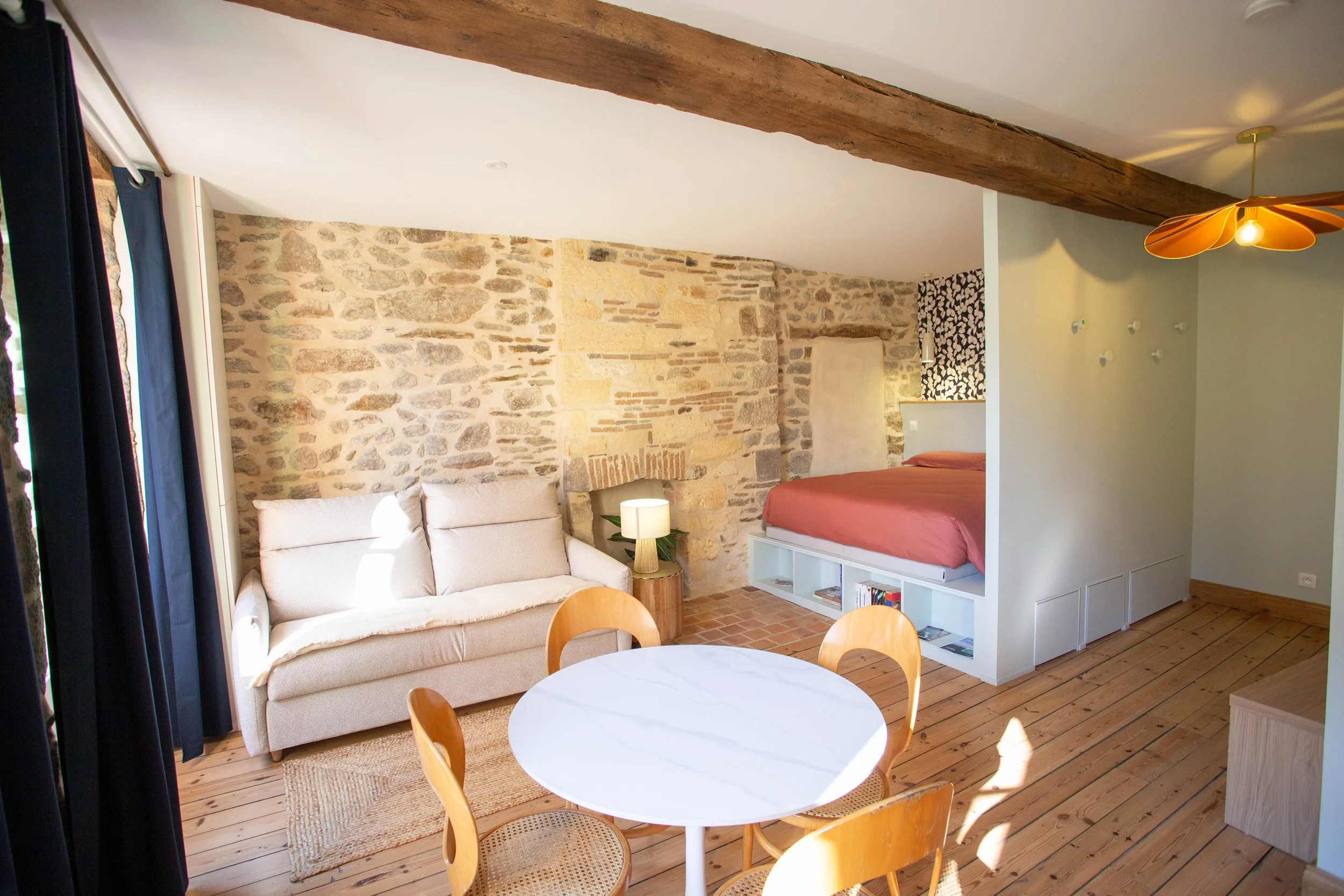 Un séjour ouvert avec un canapé beige, une table ronde blanche, des chaises en bois, un mur en pierre avec une cheminée, un lit en mezzanine avec rangement en dessous, et un sol en bois.