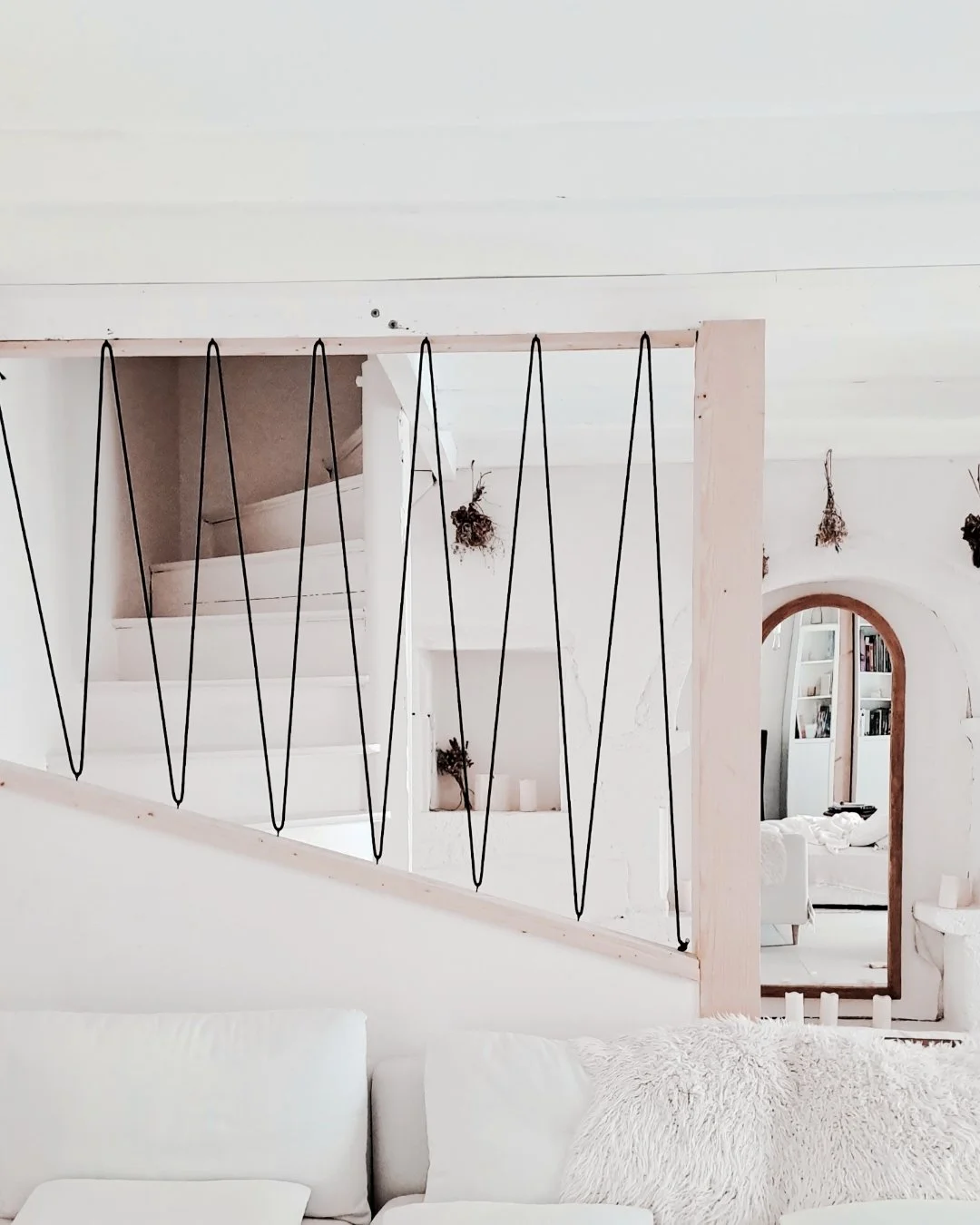 Intérieur d'une maison blanc avec une rampe en métal noir décorative séparant un salon et un escalier en ruche, avec un miroir arqué et étagères remplies de livres en arrière-plan.
