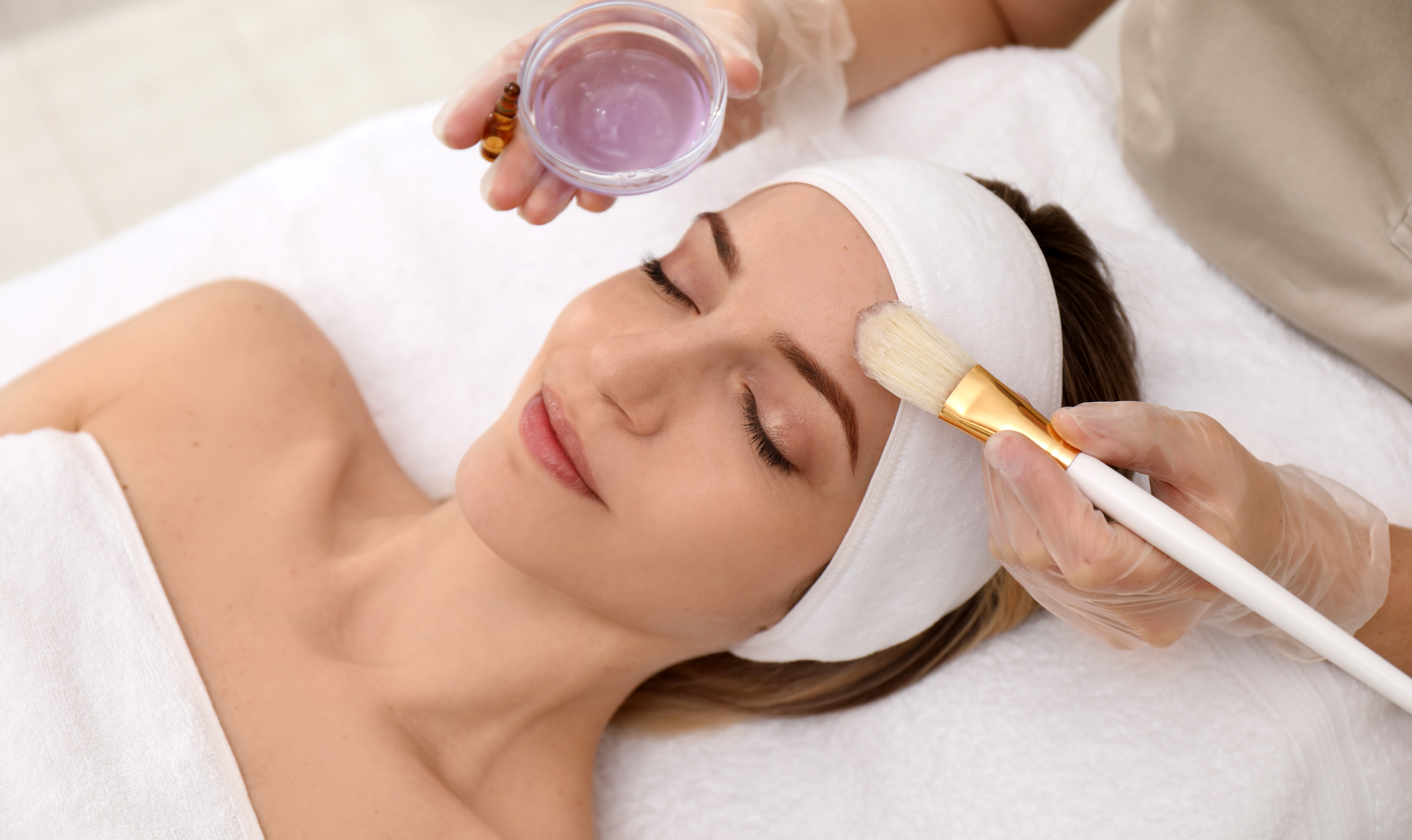 A woman receiving a facial treatment at a spa, lying down with a white towel and headband, while a skincare professional applies cream with a brush.