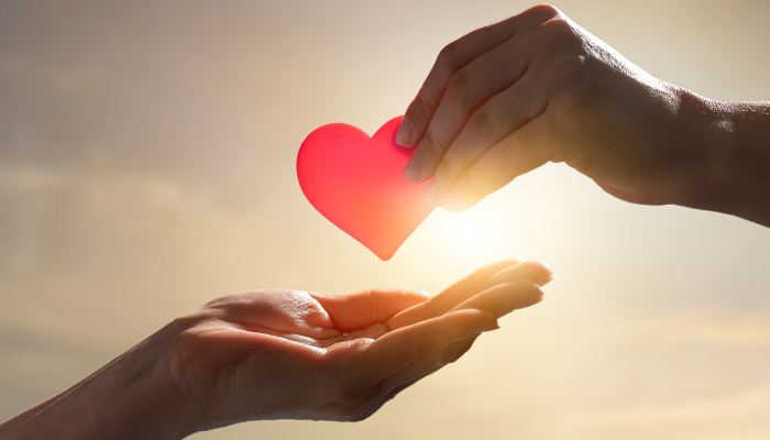 Two hands exchange a heart with the sun in the background.