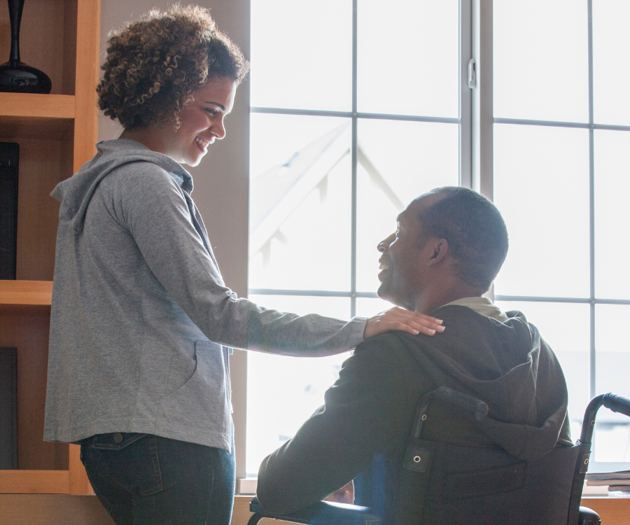 A woman stands with her hand on the shoulder of a man in a wheelchair as they look through the window in their house.