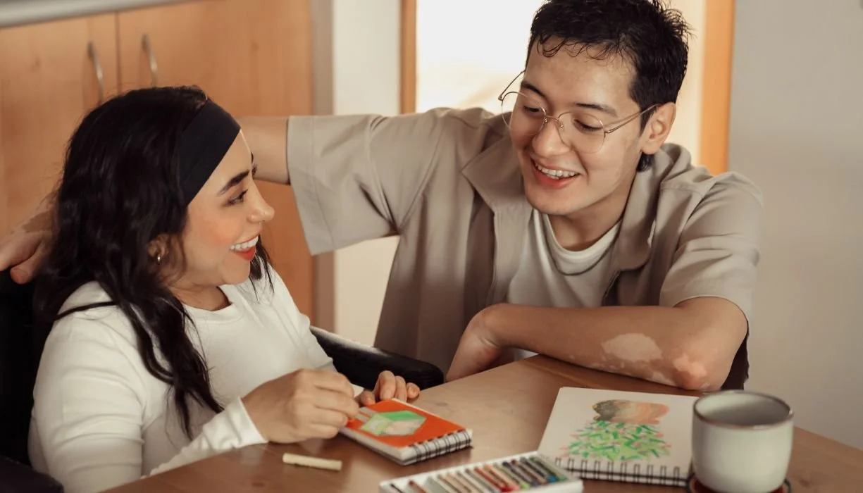A staff member smiles at a woman with disabilities as they color a piece of art in the home.
