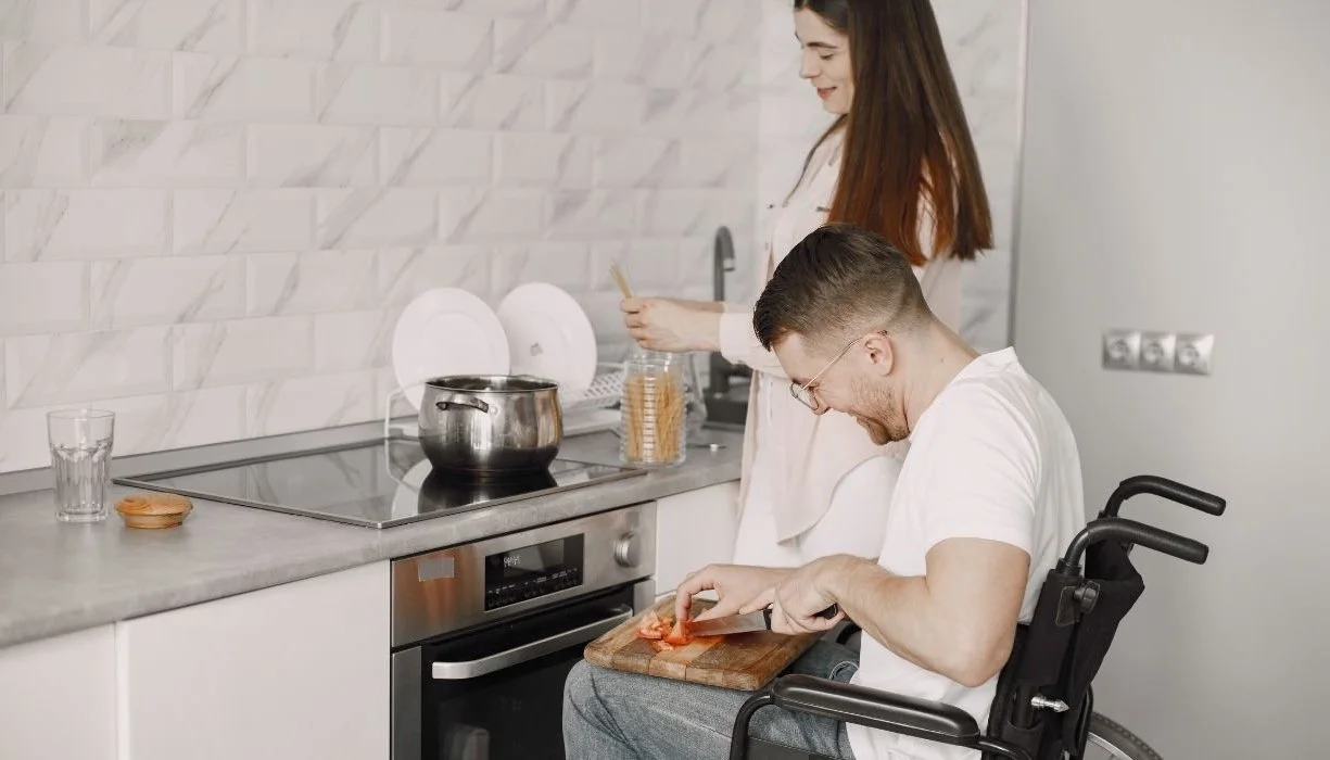 A man in a wheelchair cuts a tomato on a cutting board while a woman measures spaghetti for a pot on the stove.