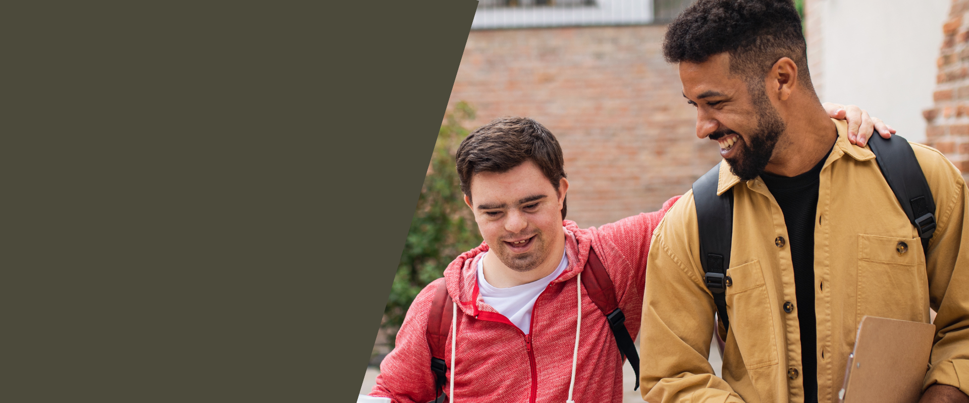 Man with down syndrome wears a red shirt and wraps his arm around another man as they walk and laugh together.