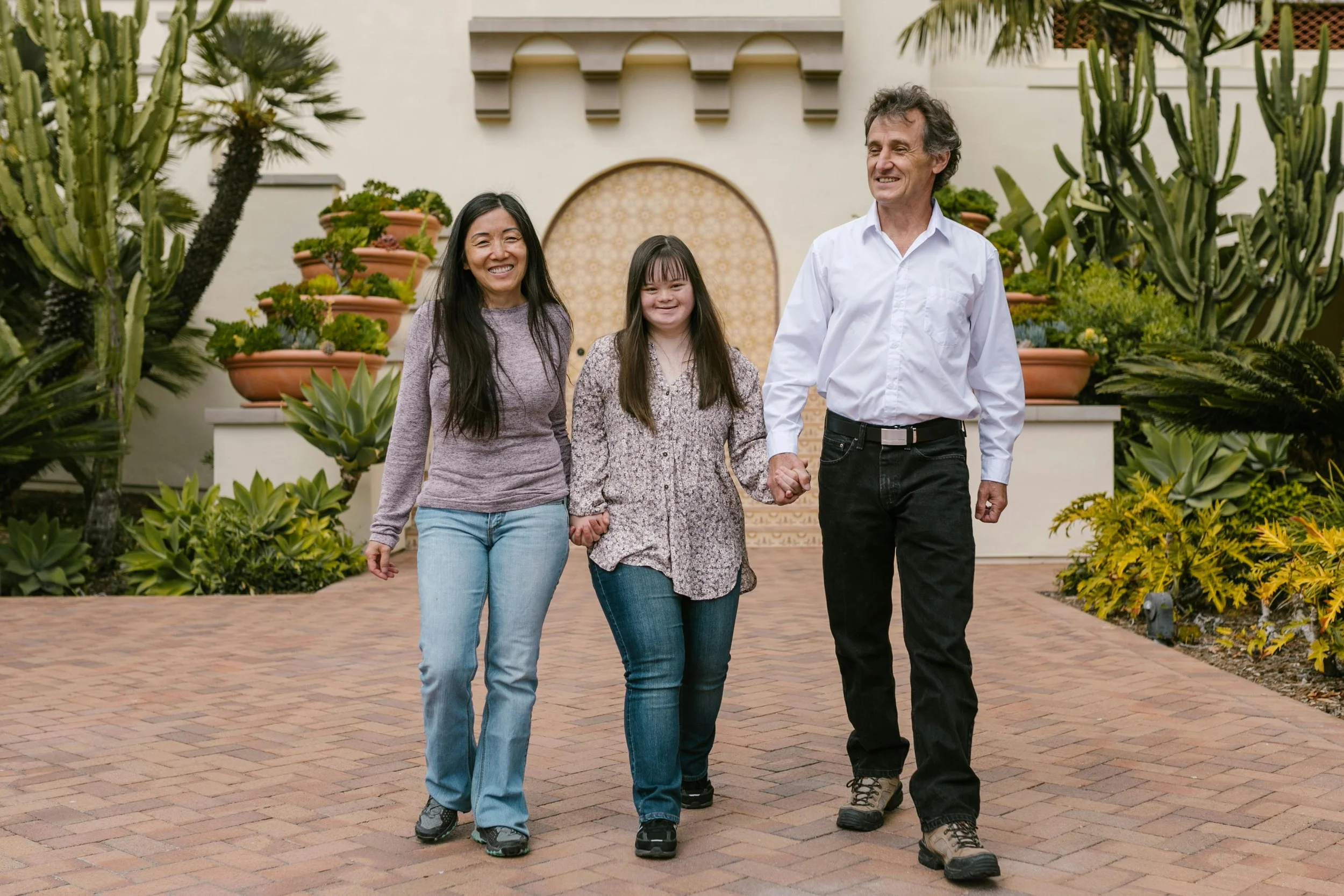 Woman with Down syndrome stands between two people and holds their hands while walking in a garden smiling.