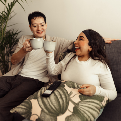 Two people sitting on a sofa, smiling and toasting mugs, with a decorative pillow and a TV remote between them.