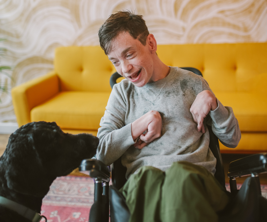 Man in a wheelchair wearing a grey sweater and green pants smiles at a black therapy dog.