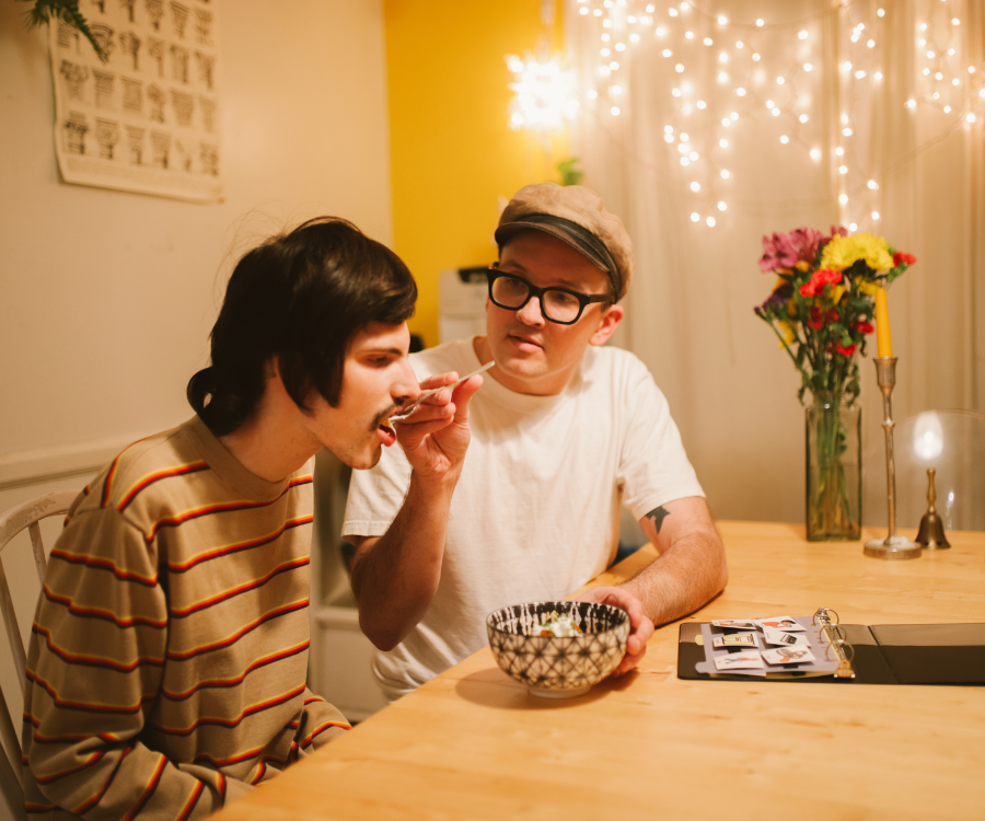 A man wearing a hat and glasses help to feed another man with disabilities wearing a striped shirt