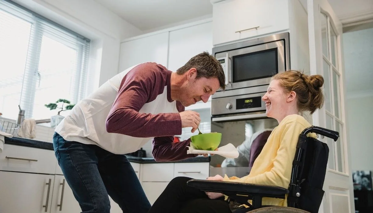A father holds a green bowl and starts to spoon some food for his daughter with disabilities while standing in their kitchen.