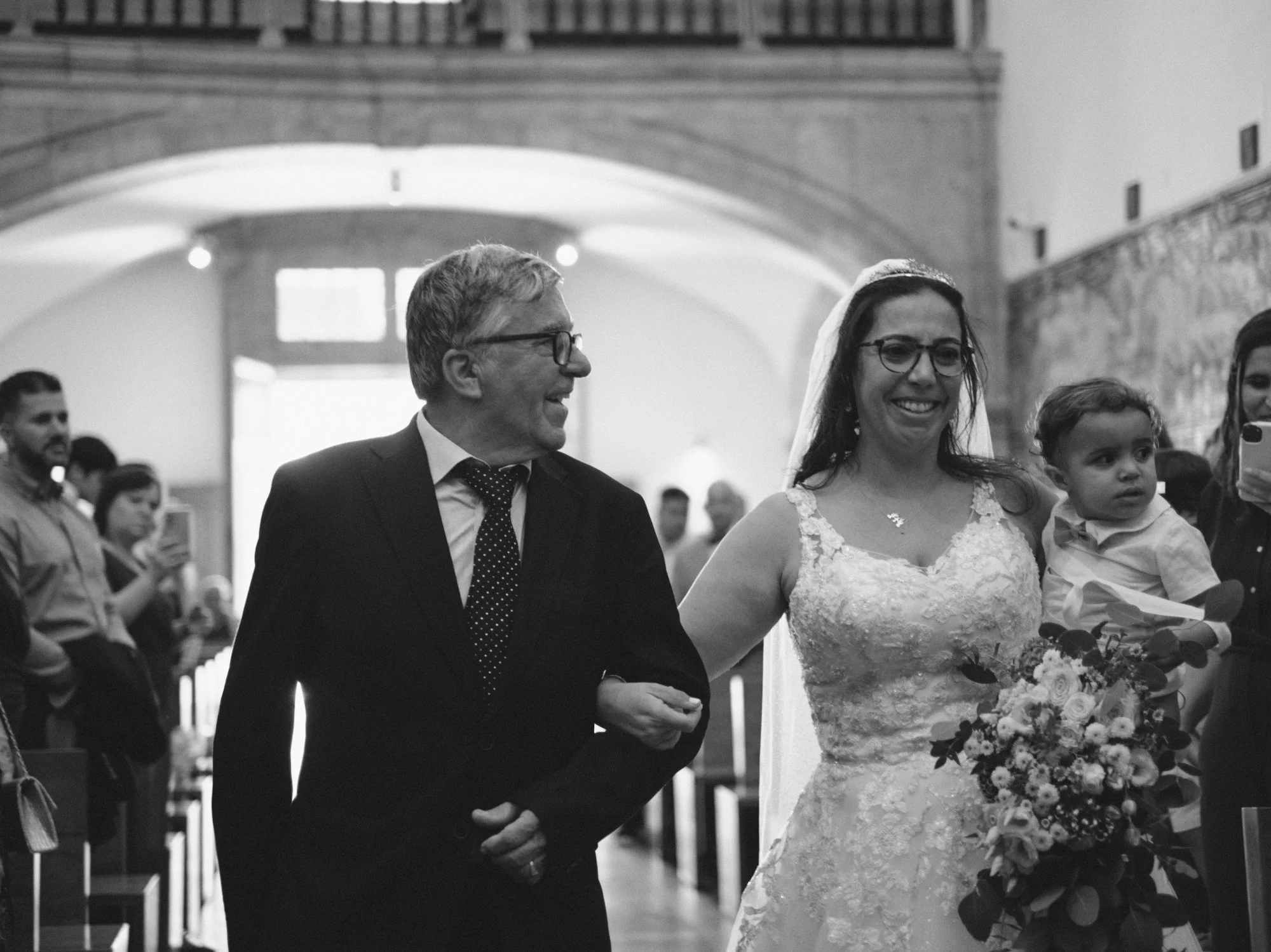 A bride walking down the aisle with her father holding her arm, carrying a bouquet of flowers, smiling, in a wedding ceremony inside a church.