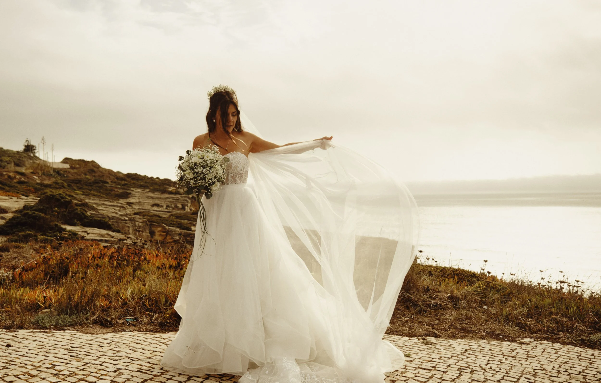 Noiva em vestido branco a segurar bouquet junto à costa ao pôr do sol numa sessão de fotografia de casamento em Lisboa