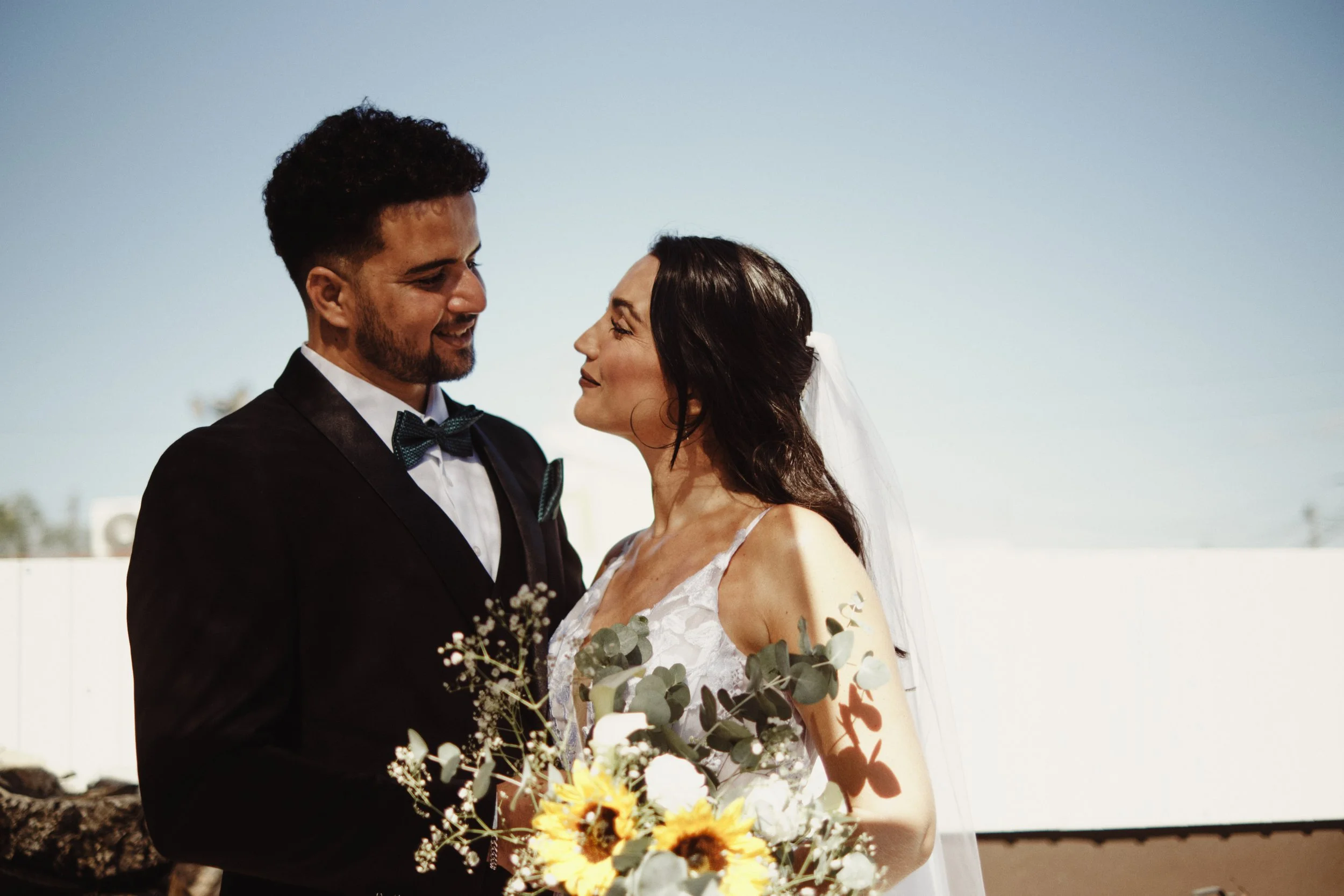 Noivos frente a frente numa sessão de fotografia de casamento ao ar livre, com bouquet de girassóis e céu azul, fotografia de casamento profissional