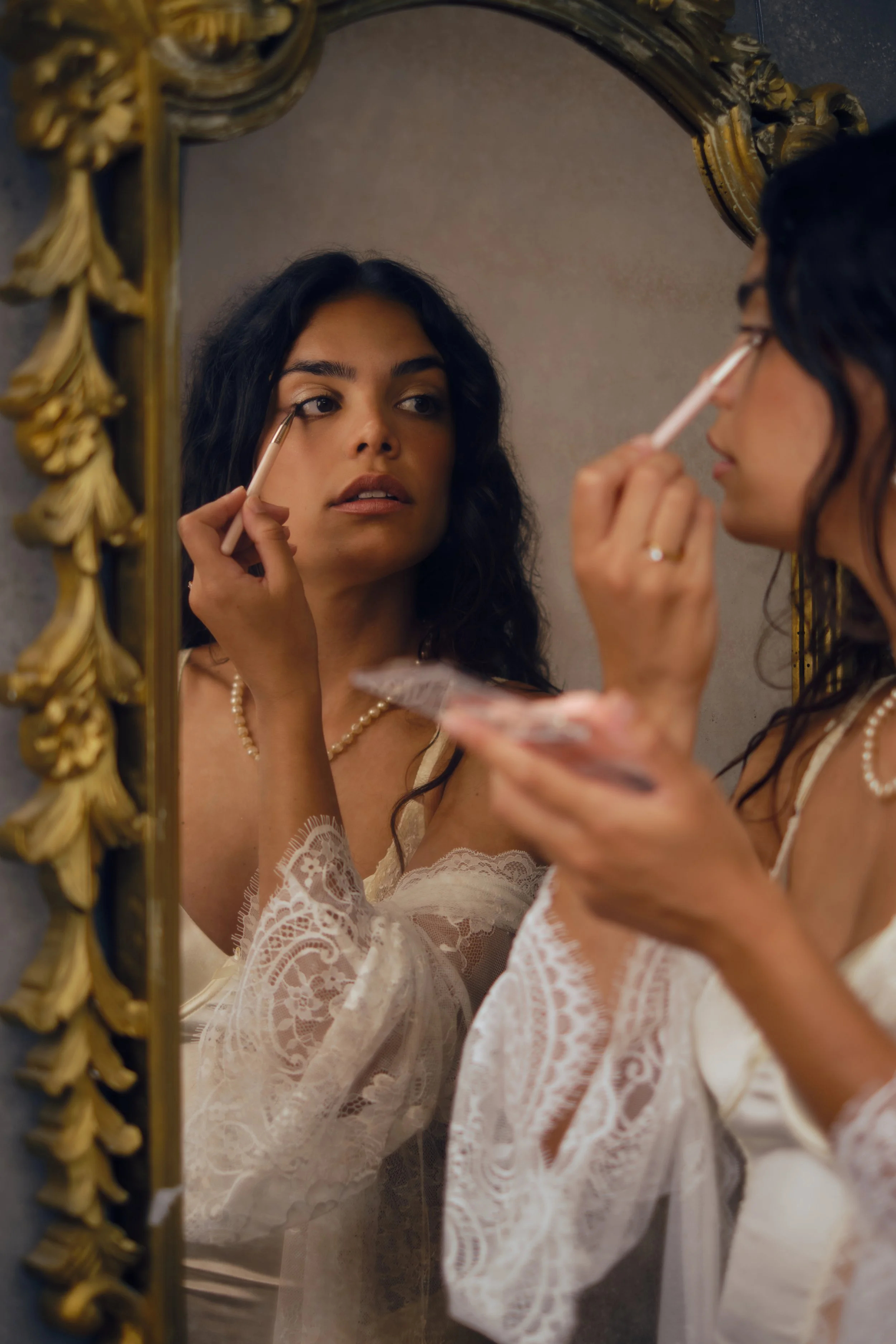 Mulher com cabelo ondulado a maquilhar-se em frente a espelho dourado numa sessão fotográfica feminina em Lisboa, com colar de pérolas e roupa rendada