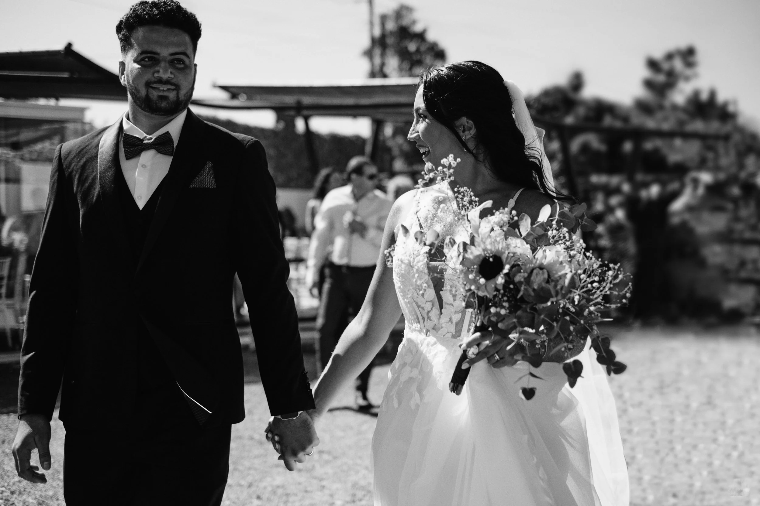 Casal recém-casado a caminhar de mãos dadas ao ar livre em Lisboa, noiva com bouquet e noivo de fato e gravata borboleta, fotografia de casamento em preto e branco