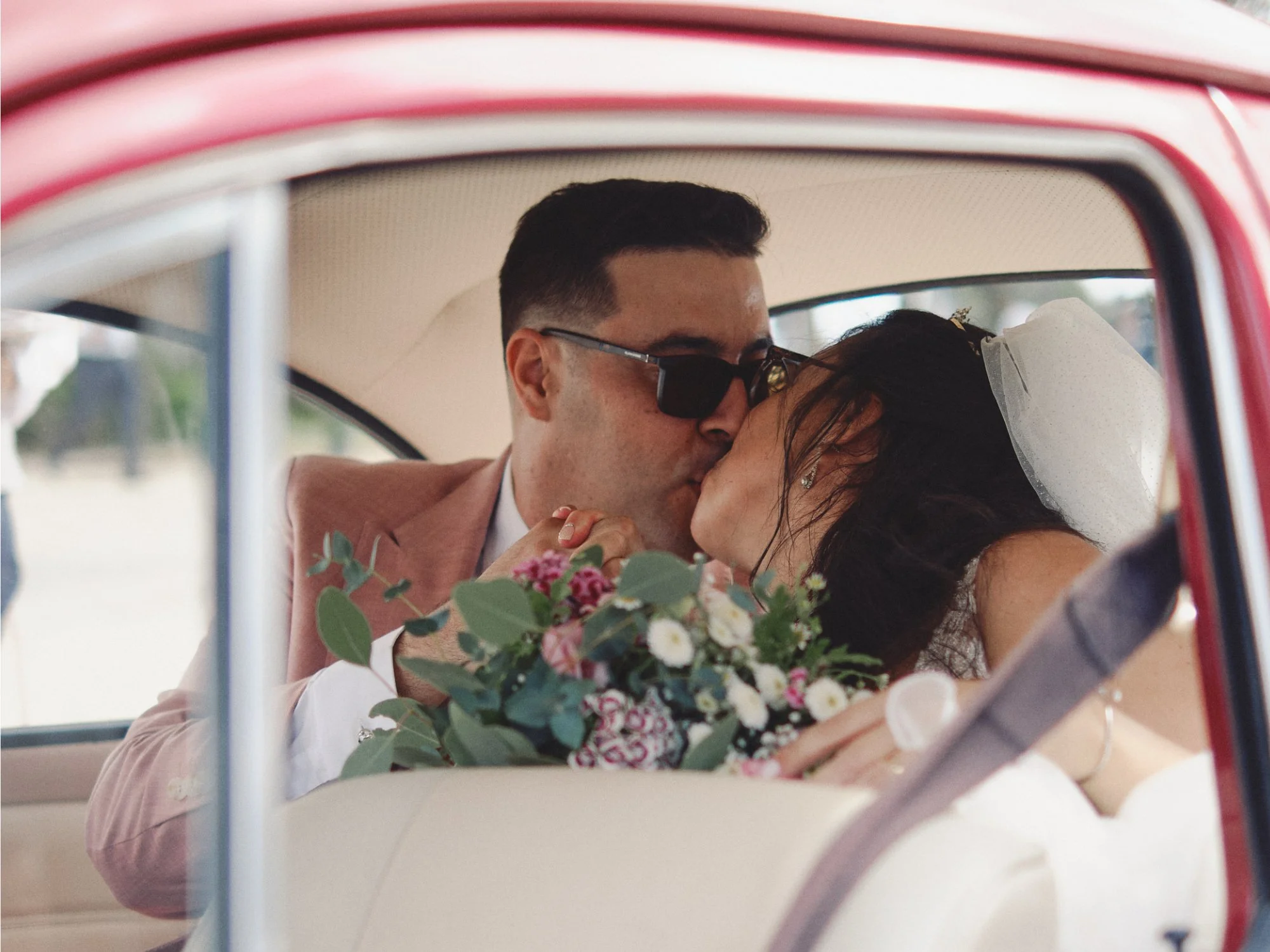 A newlywed couple sharing a kiss inside a vintage car. The groom is wearing sunglasses and a pink suit, while the bride has a veil and is holding a bouquet of flowers.