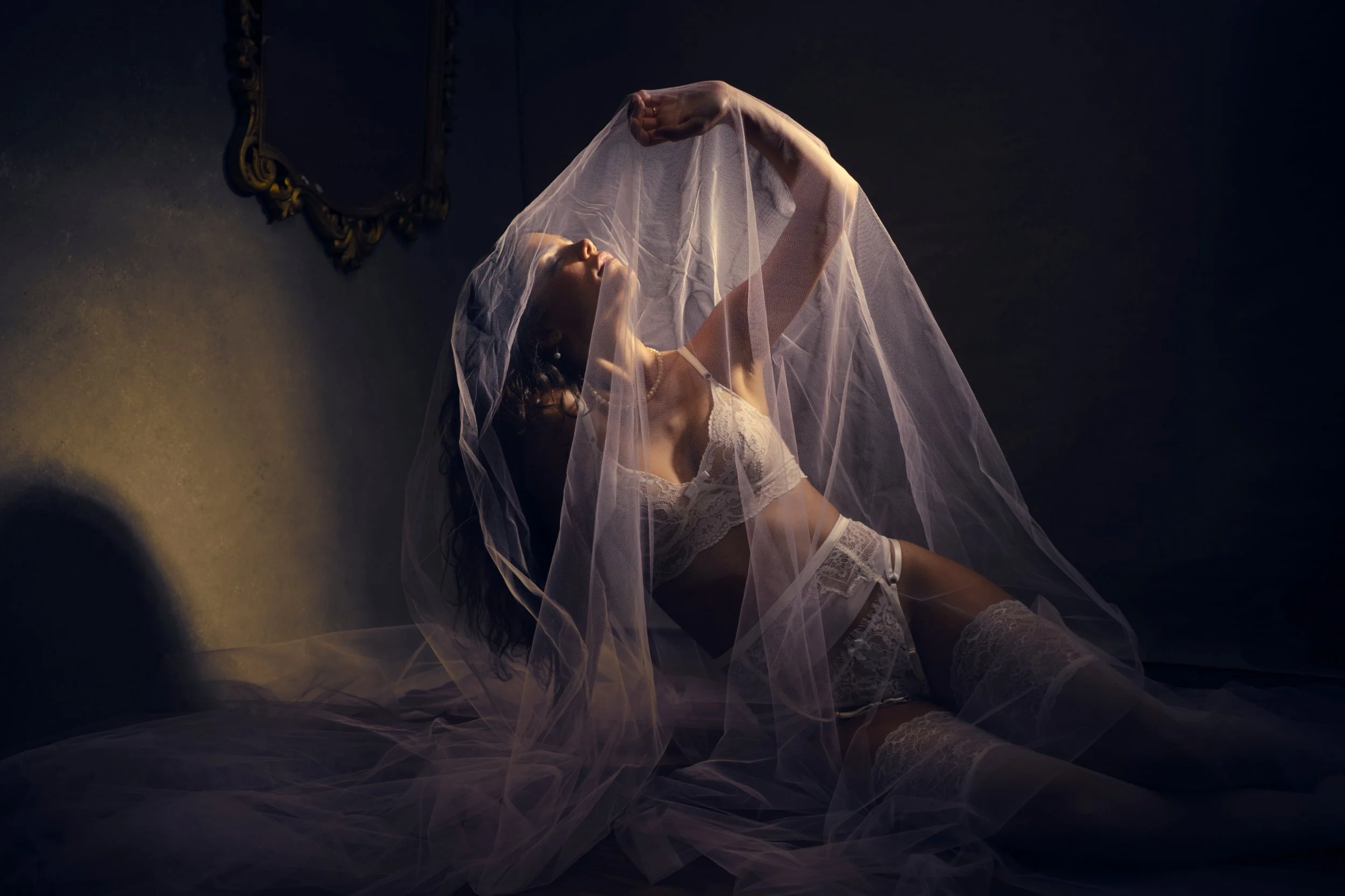 Woman in white lingerie and stockings, sitting on the floor, partially covered by a sheer veil, with a decorative mirror in the background, in a dimly lit room.