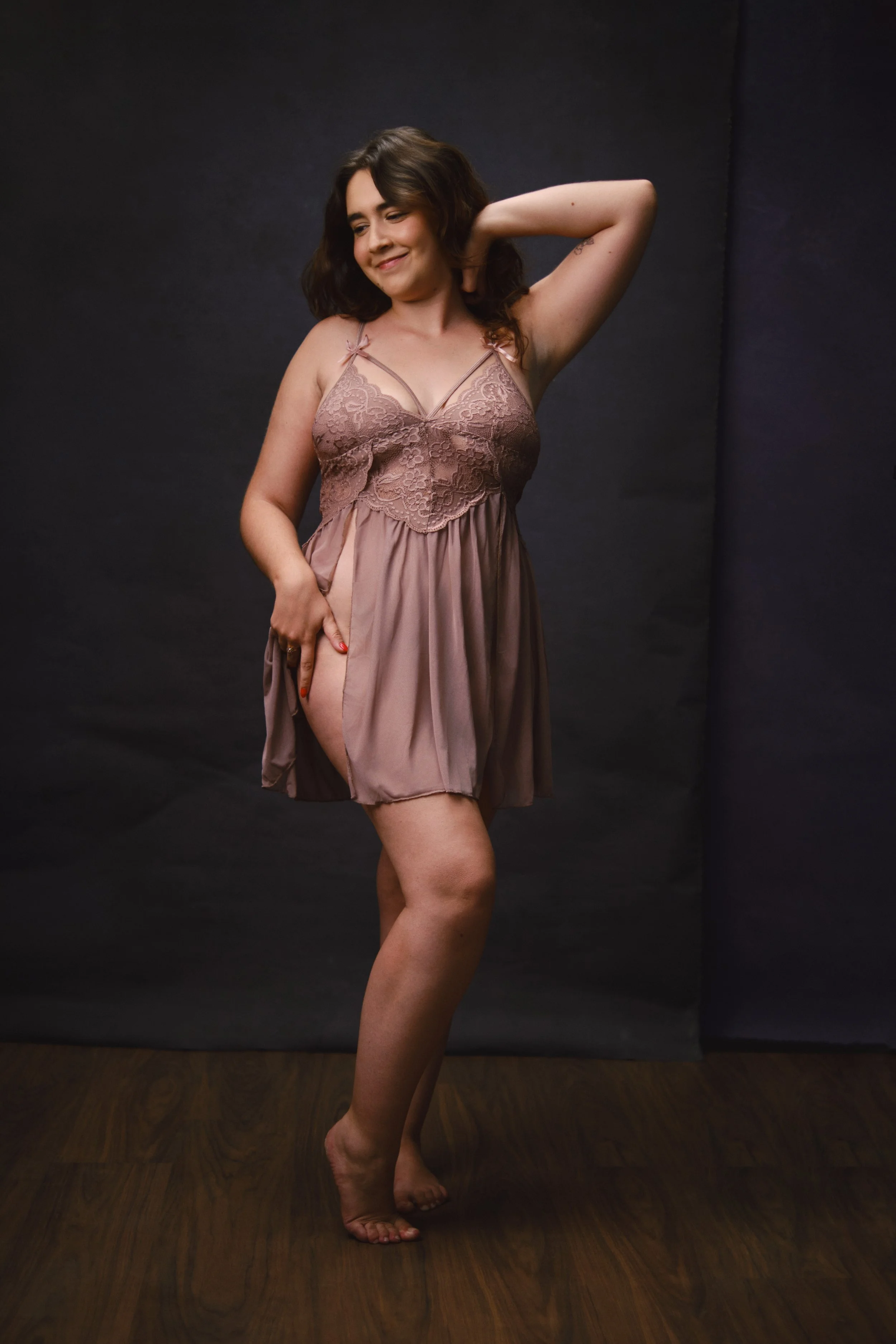 A woman is standing in front of a dark backdrop, smiling with one eye closed. She is wearing a beige lace and satin slip dress, with one hand behind her head and the other pulling up the dress to reveal her thigh, standing on a wooden floor.
