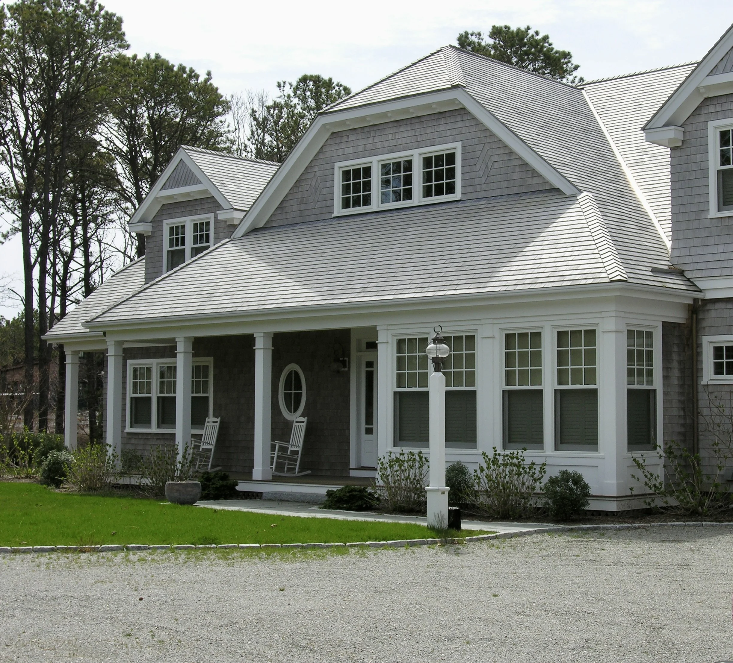 A two-story gray house with a front porch, several white-framed windows, and a sloped roof, surrounded by trees and a gravel driveway.