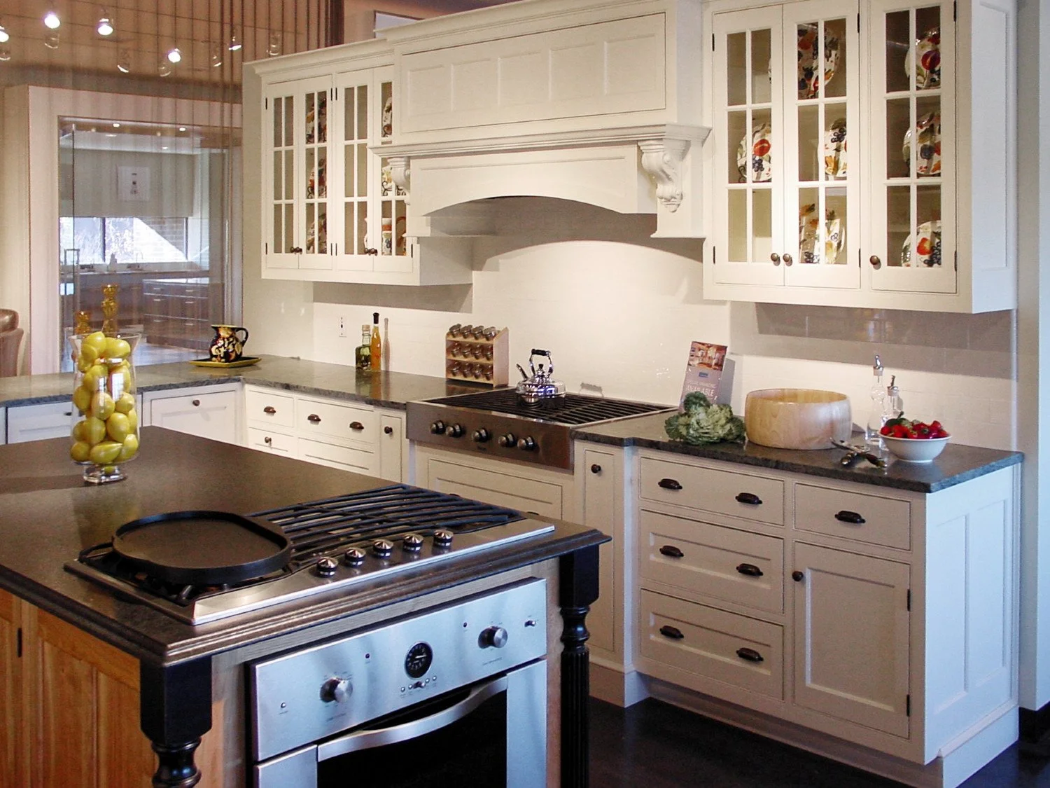 A kitchen with white cabinets, a black countertop, and stainless steel appliances, including a stove and oven. There are decorative items and food on the countertops, and a sliding door leading to an outdoor space.