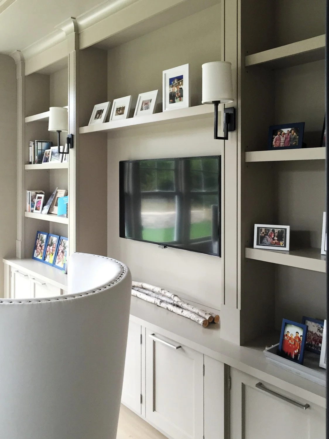 Living room built-in white cabinetry with a mounted flat-screen tv, picture frames, and decorative logs, a white armchair in the foreground, and black wall sconces with white shades