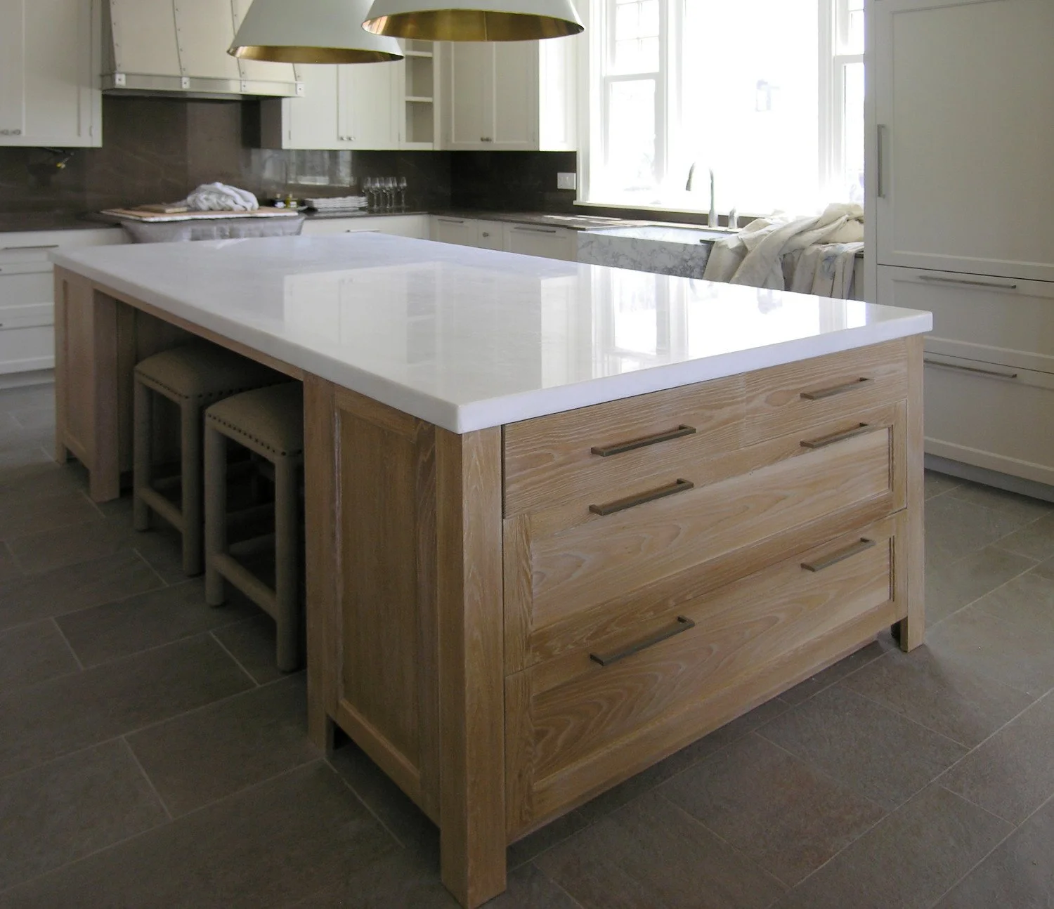 Modern kitchen with a large island featuring a white countertop and wooden drawers, with three stools tucked underneath. In the background, white cabinets, a window, and hanging gold light fixtures are visible.