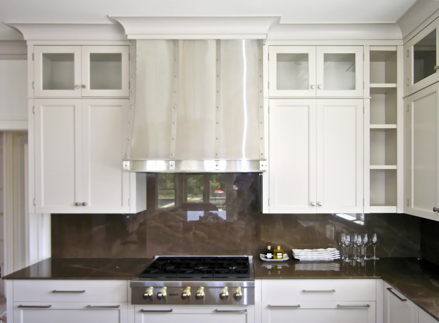 White kitchen cabinetry with glass-front upper cabinets, a stainless steel range hood, a dark countertop, a gas stove, and a brown marble backsplash. There are wine glasses, plates, and condiment bottles on the counter.